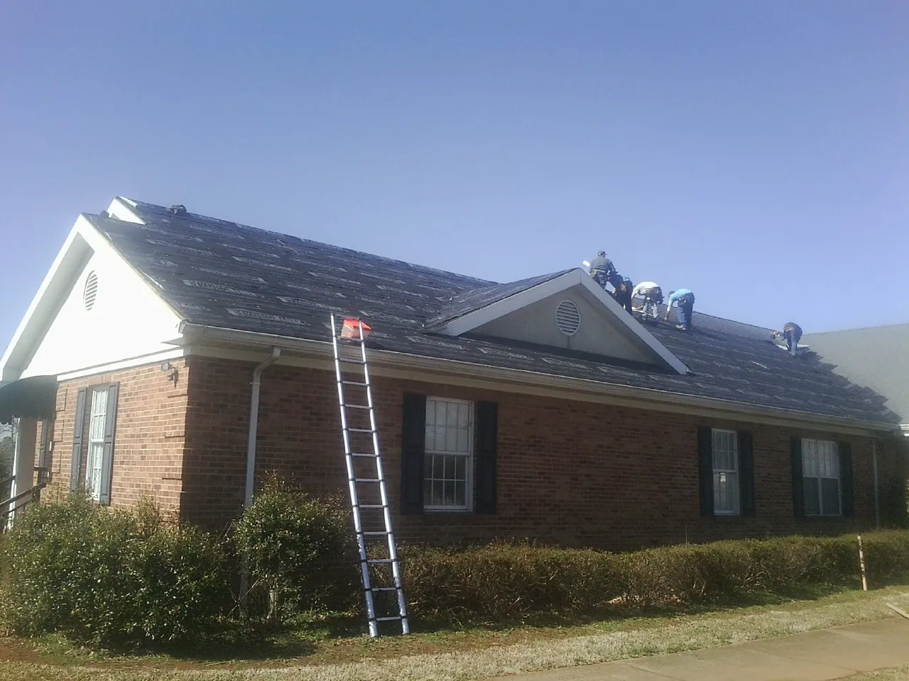 Crew tearing shingles off a brick ranch roof with a ladder set — Emergency Roof Repair work by our crew in Iberia