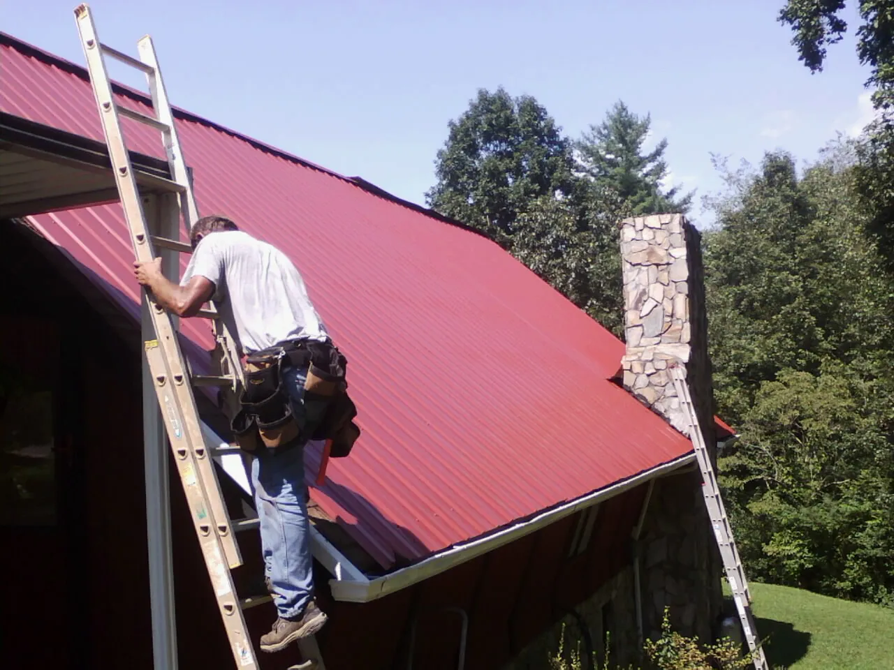 Roofer on a ladder beside a red metal roof and stone chimney — Soffit & Fascia Repair by Roofing Brothers in Iberia